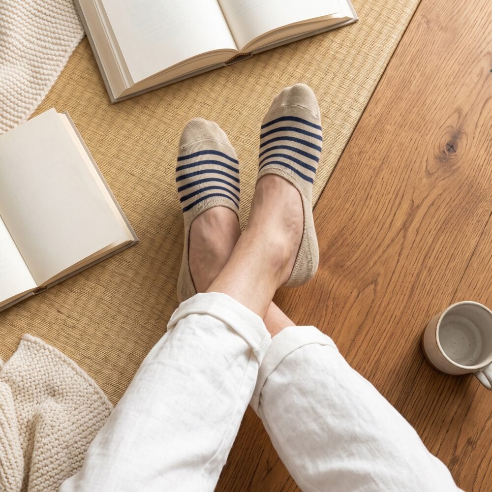 A close-up of a person's feet wearing low-cut beige no show socks with navy blue stripes across the top, standing on a polished hardwood floor.