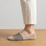 A close-up of a person's feet wearing low-cut beige no show socks with navy blue stripes across the top, standing on a polished hardwood floor.