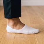 Person's foot wearing white breathable no show socks on a wooden floor, paired with dark trousers.