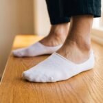Person's foot wearing white breathable no show socks on a wooden floor, paired with dark trousers.