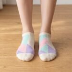 A close-up of a person's feet wearing ultra-low cut no show socks with a pastel pink, lavender, and mint green geometric pattern, standing tiptoe on a wooden surface.
