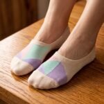 A close-up of a person's feet wearing ultra-low cut no show socks with a pastel pink, lavender, and mint green geometric pattern, standing tiptoe on a wooden surface.