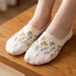A close-up of a person's feet wearing cream-colored no show socks with a delicate yellow wildflower pattern, standing on a light wood floor.