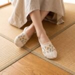 A close-up of a person's feet wearing cream-colored no show socks with a delicate yellow wildflower pattern, standing on a light wood floor.