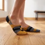 A close-up side view of a person's feet wearing ultra-low cut no show socks with a mustard yellow, black, and grey geometric pattern, standing on a sunlit wooden floor.