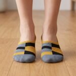 A close-up side view of a person's feet wearing ultra-low cut no show socks with a mustard yellow, black, and grey geometric pattern, standing on a sunlit wooden floor.