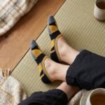 A close-up side view of a person's feet wearing ultra-low cut no show socks with a mustard yellow, black, and grey geometric pattern, standing on a sunlit wooden floor.