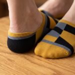 A close-up side view of a person's feet wearing ultra-low cut no show socks with a mustard yellow, black, and grey geometric pattern, standing on a sunlit wooden floor.
