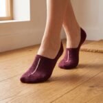 A close-up of a person's feet wearing deep burgundy no show socks with a single vertical white stripe, standing on a light wood floor.