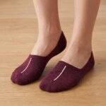 A close-up of a person's feet wearing deep burgundy no show socks with a single vertical white stripe, standing on a light wood floor.