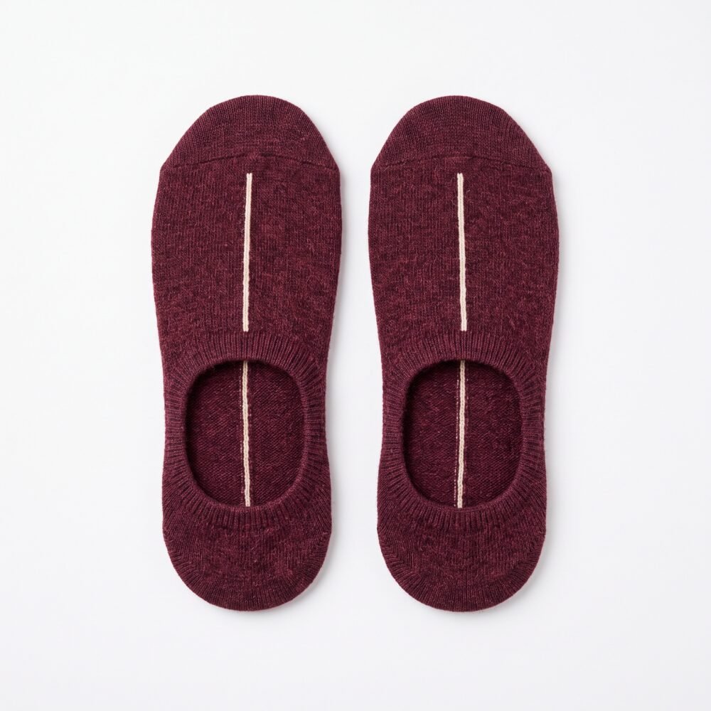 A close-up of a person's feet wearing deep burgundy no show socks with a single vertical white stripe, standing on a light wood floor.