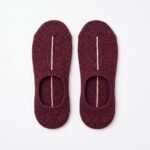 A close-up of a person's feet wearing deep burgundy no show socks with a single vertical white stripe, standing on a light wood floor.