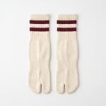 A pair of cream-colored crew-length Tabi socks featuring a split toe and two maroon stripes at the ribbed cuff, standing upright on a sunlit wooden floor.
