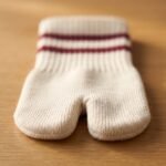 A pair of cream-colored crew-length Tabi socks featuring a split toe and two maroon stripes at the ribbed cuff, standing upright on a sunlit wooden floor.