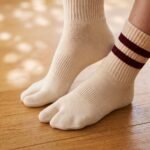 A pair of cream-colored crew-length Tabi socks featuring a split toe and two maroon stripes at the ribbed cuff, standing upright on a sunlit wooden floor.