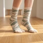 A pair of tall cream Tabi socks featuring an abstract sage green wavy pattern, laid flat against a crisp white background.
