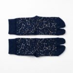 A person wearing navy blue crew-length Tabi socks featuring a gold and white star constellation pattern, standing on a light wood floor.