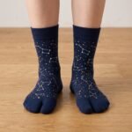 A person wearing navy blue crew-length Tabi socks featuring a gold and white star constellation pattern, standing on a light wood floor.