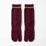 A person wearing heathered burgundy ankle-length Tabi socks with a single thin white stripe at the ribbed cuff, standing on a light wood floor.