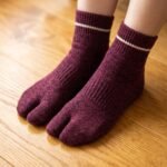 A person wearing heathered burgundy ankle-length Tabi socks with a single thin white stripe at the ribbed cuff, standing on a light wood floor.