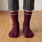 A person wearing heathered burgundy ankle-length Tabi socks with a single thin white stripe at the ribbed cuff, standing on a light wood floor.