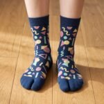 A person wearing navy blue crew-length Tabi socks featuring a colorful retro 80s geometric Memphis pattern, standing on a sunlit light wooden floor.