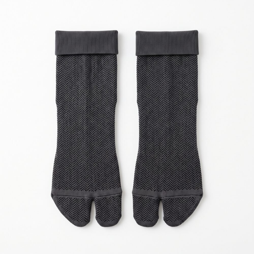 A person wearing charcoal grey ankle-length Tabi socks with a herringbone knit texture and folded cuffs, standing on a sunlit wooden floor.