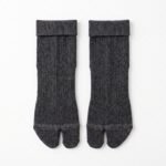 A person wearing charcoal grey ankle-length Tabi socks with a herringbone knit texture and folded cuffs, standing on a sunlit wooden floor.