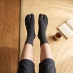 A person wearing charcoal grey ankle-length Tabi socks with a herringbone knit texture and folded cuffs, standing on a sunlit wooden floor.
