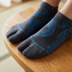 A pair of dark grey crew-length Tabi socks featuring a bright blue abstract wireframe pattern, laid flat against a crisp white background.