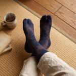 A person wearing navy blue crew-length Tabi socks featuring a copper geometric line pattern, standing on a light wood floor.
