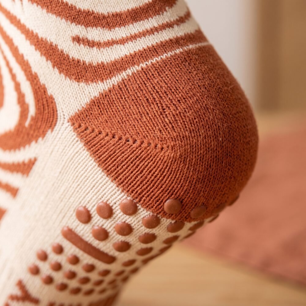 Close-up of a woman's foot wearing a tan and brown swirl pattern crew grip sock, highlighting the rust-colored silicone non-slip dots and bars on the heel and sole.