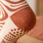 Close-up of a woman's foot wearing a tan and brown swirl pattern crew grip sock, highlighting the rust-colored silicone non-slip dots and bars on the heel and sole.